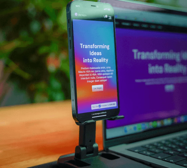 A smartphone is mounted on a stand, displaying a website with colorful graphics and the text 'Transforming Ideas into Reality'. Beside it, a laptop screen shows a blurred version of the same website. The background is soft and out of focus, with green foliage adding a hint of nature.